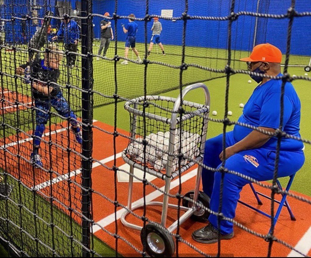 Coach Noochie Varner sitting behind the screen feeding batting practice to a young hitter in the indoor cage