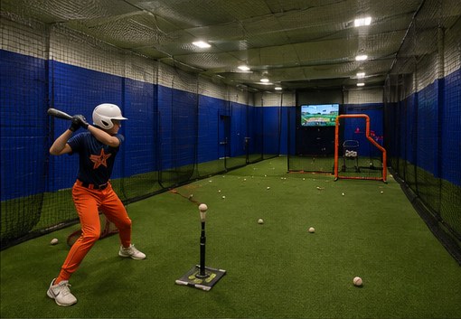 Player working off a batting tee in the indoor cage with the HitTrax monitor visible on the wall